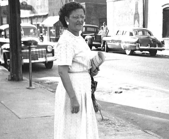 Louise Wingate Beard standing on a downtown sidewalk holding a rolled newspaper, 1940s cars behind her